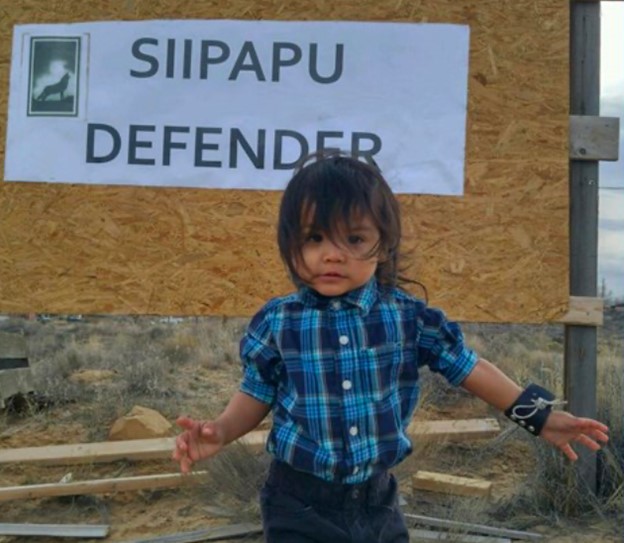 Young Hopi child standing in front of a sign that reads "Siipapu Defender"