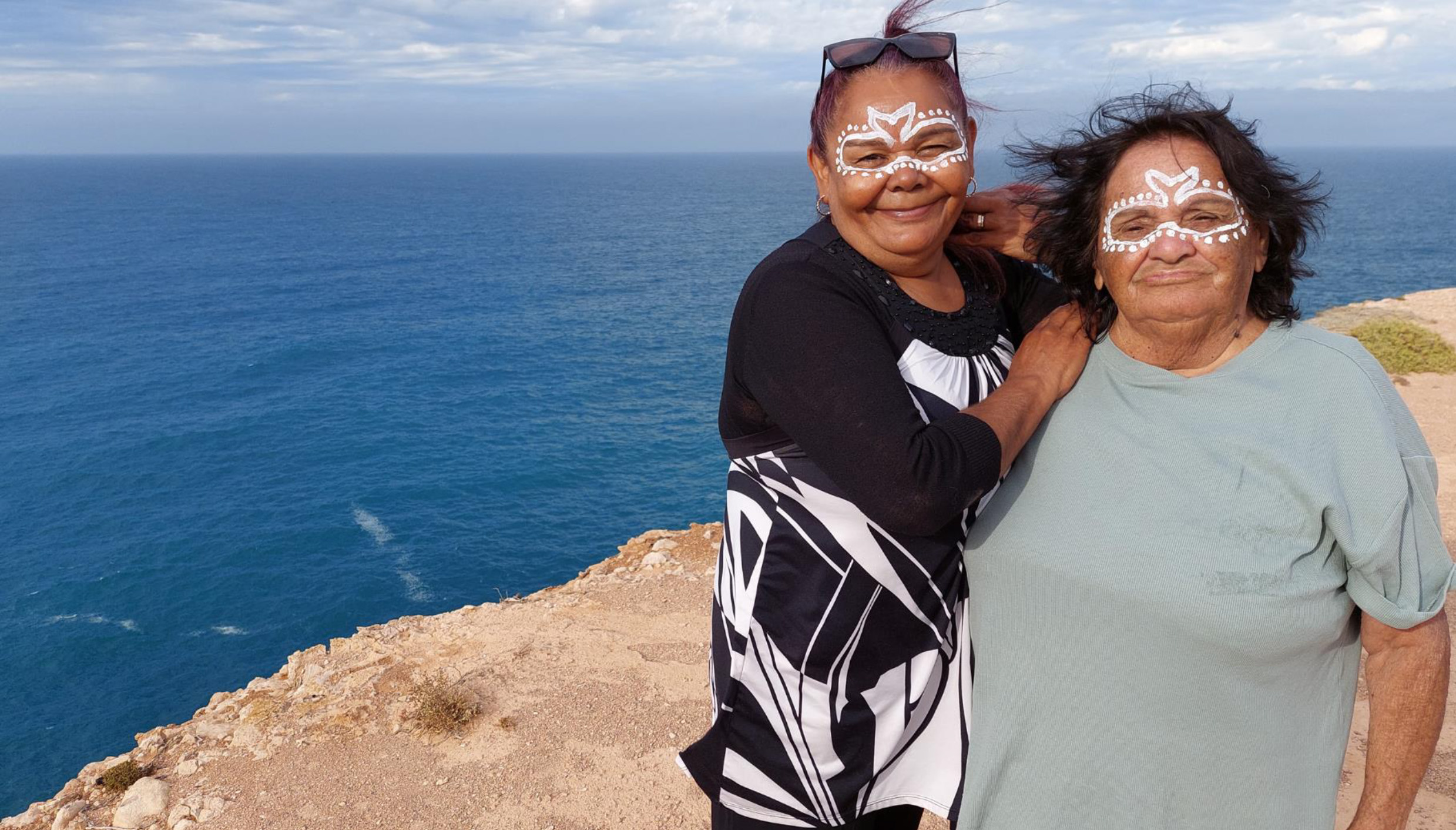 Photo of two Aboriginal women standing atop a cliff. Their faces are painted with traditional Mirning symbols and you can see the ocean spanning far and wide behind them.
