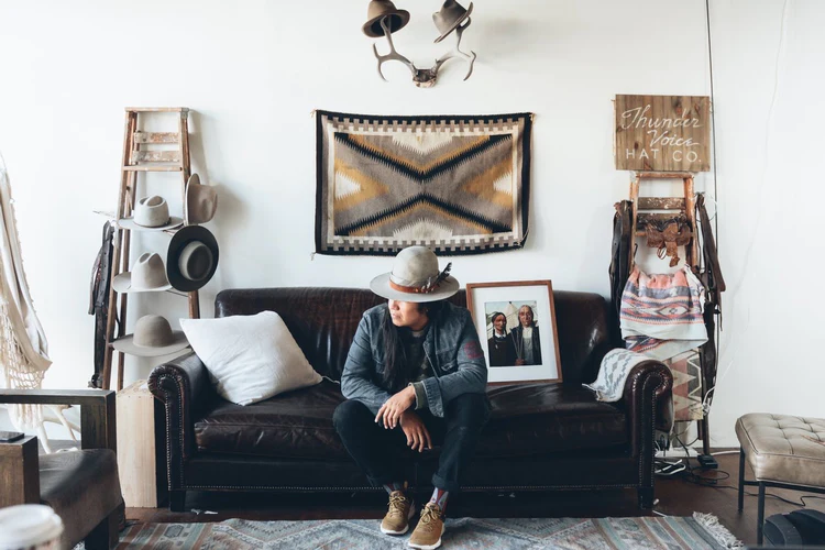 Photo of a man sitting on a couch surrounded by Indigenous art. He is wearing a Thunder Voice Hat. 